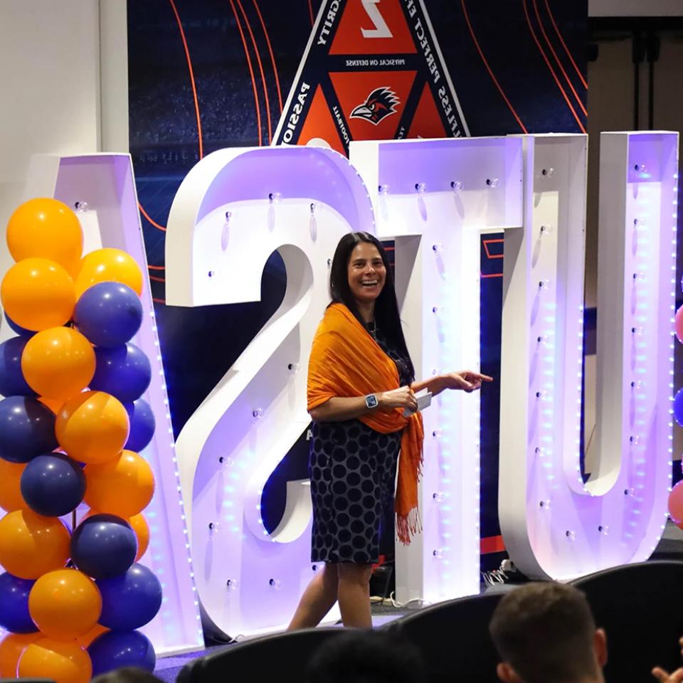 世界杯官方app Vice President for Intercollegiate Athletics Lisa Campos stands smiling next to large, illuminated letters spelling '世界杯官方app', decorated with orange and blue balloons